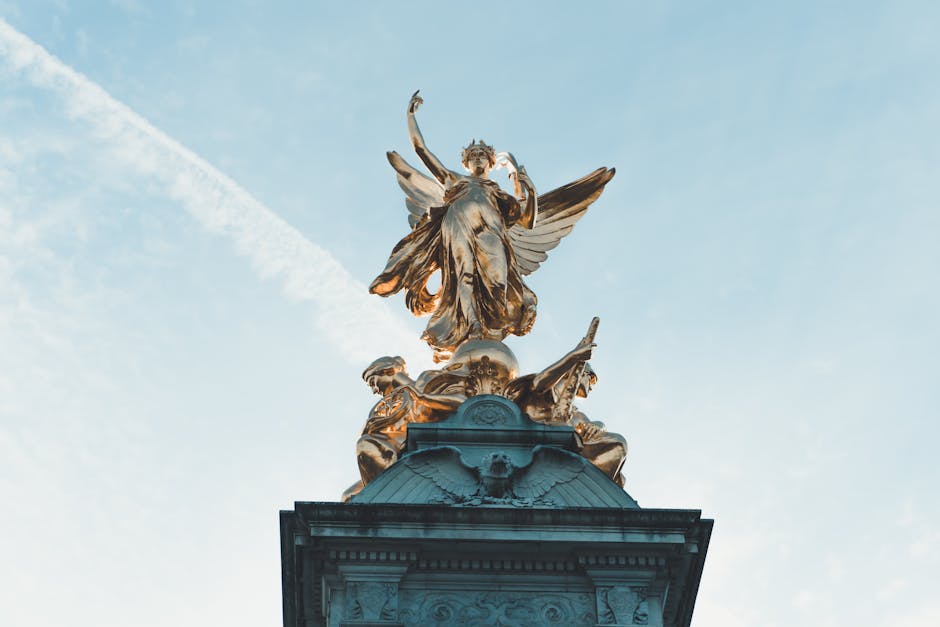 A detailed, downward-looking view of a gilded, ornate statue atop a stone pedestal, featuring an angelic figure with outstretched wings and a flowing robe, holding an object in its hand. Surrounding t