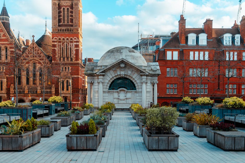 A rooftop garden situated in an urban setting with a paved walking area leading to a small domed structure made of white stone, featuring decorative architectural elements and a dark-green metal raili