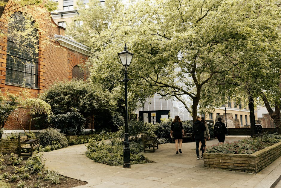A rooftop garden situated in an urban setting with a paved walking area leading to a small domed structure made of white stone, featuring decorative architectural elements and a dark-green metal raili
